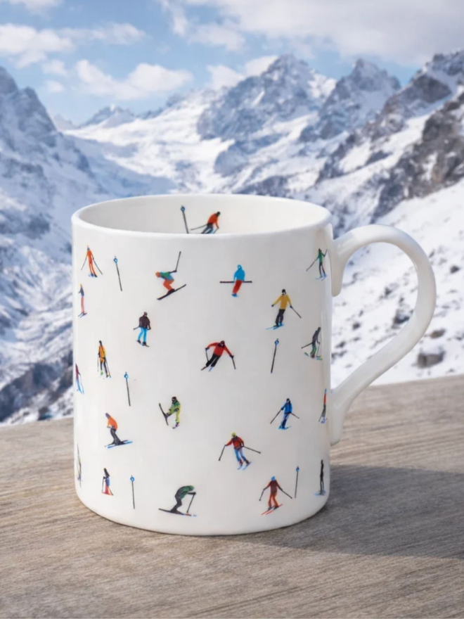 Ski Slope Mug. A white mug patterned with a simple multicoloured skier design, set against a mountain scene.