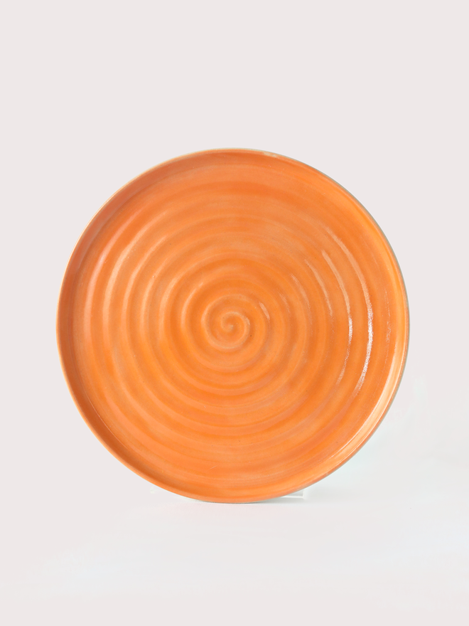 A handmade ceramic lunch plate in glossy mandarin orange glaze, photographed from above against a plain light grey background, showing a natural concentric swirl texture from the wheel-throwing process