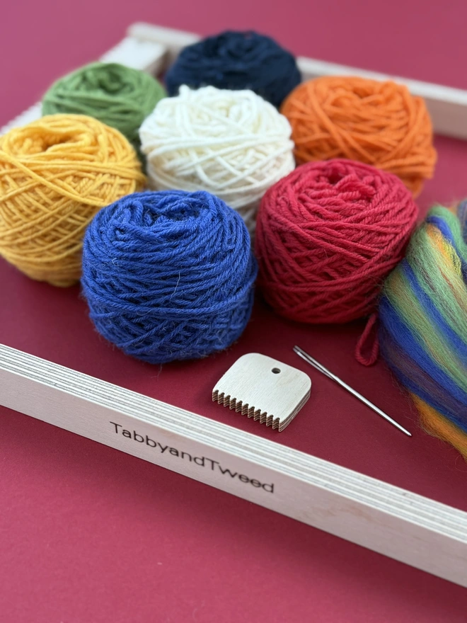 Wooden beige frame loom with seven balls of yarn in white, green, blue, orange, yellow, red and cream plus some rainbow merino fibres, a wooden comb and needle