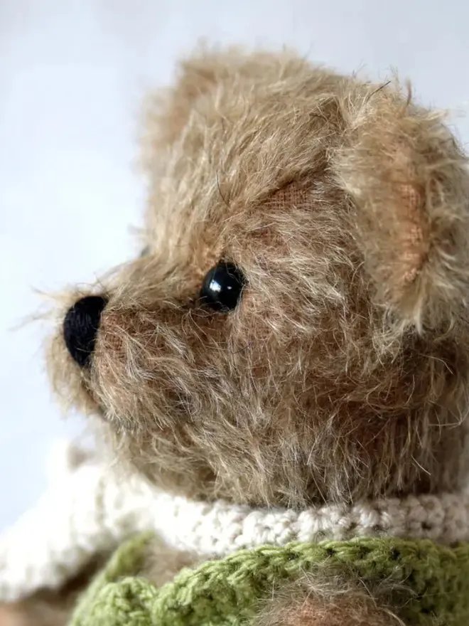Handmade brown teddy bear with a white scarf wearing a green cardigan, sitting against a plain background.