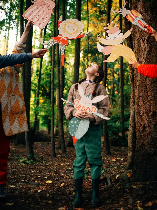 Extra Large Lion Boy Wall Puppet. a group of children in a woodland setting hold up an array of matching wooden puppets, including the lion boy puppet with its cream and gold mane and red and green checked outfit, alongside a fox, two deer figures and a hare in coordinating pink, yellow and red patterned costumes. a young girl in a knitted jumper and green trousers stands in the centre looking up at the puppets held aloft around her. the autumnal forest backdrop and playful group scene give the puppets a whimsical, storybook charm.