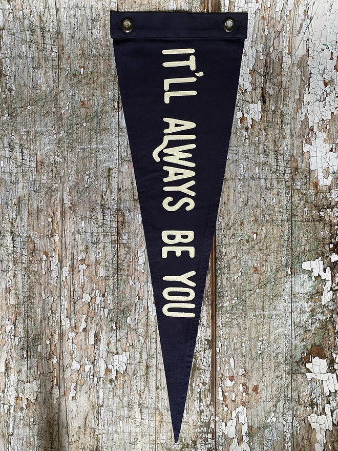 A navy pennant flag hung vertically on a wall. Written in ivory canvas are the words It'll always be you.