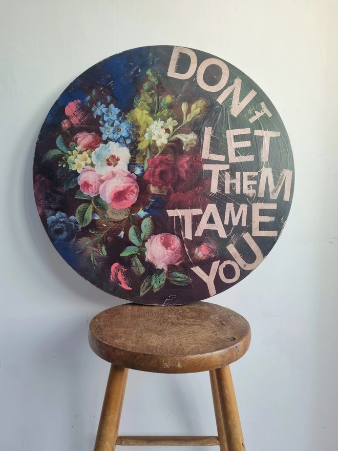 Art Panel made from a round table top. A dark background with multicolour flowers and writing in pink that says 'don't let them tame you'.