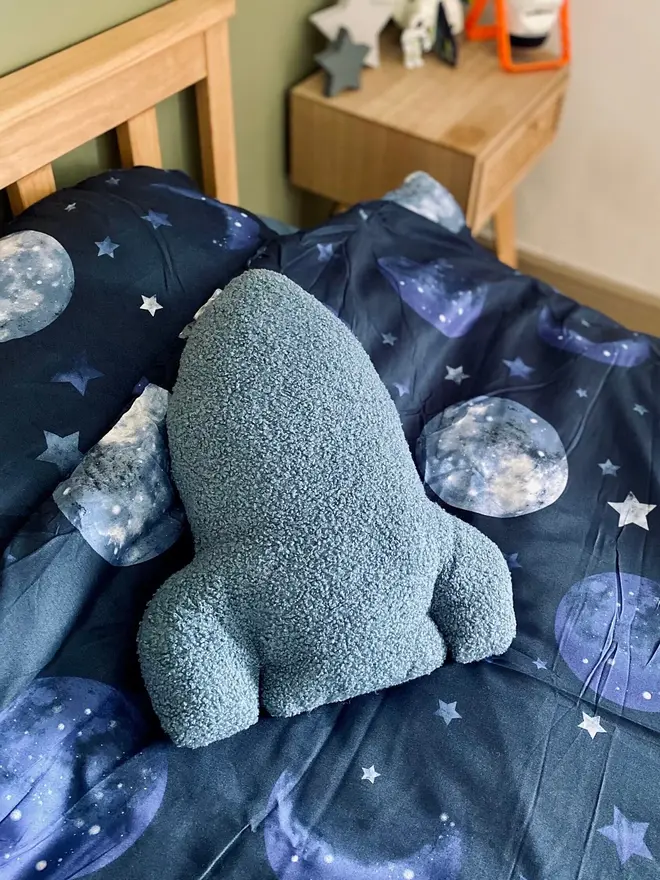 boucle rocket pillow. a blue rocket cushion on a bed with space-themed sheets.