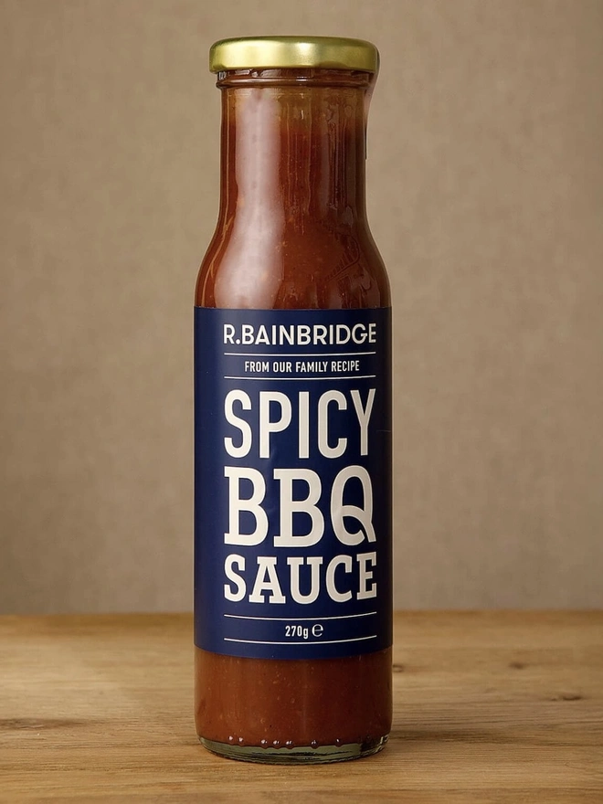 Spicy BBQ Sauce, a jar of BBQ sauce sitting on a wooden surface against a plain backdrop. 