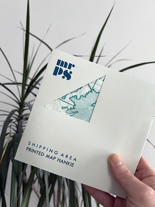 A white hand holds a Mr.PS Shipping Area Handkerchief in a packet in front of a plant