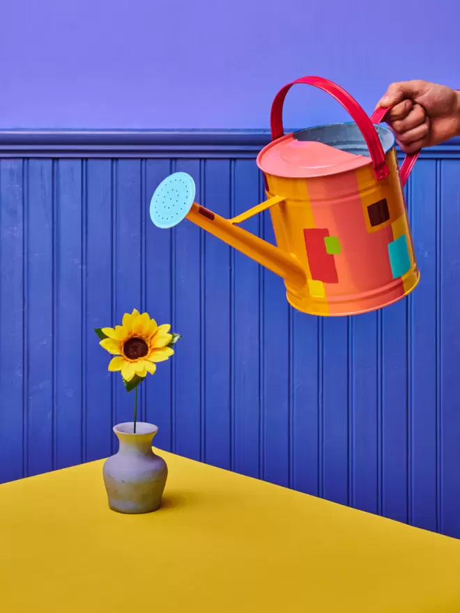 Hand painted watering can by london artist Julie-Anne, in Peach. The watering can is watering a sunflower on a yellow table.
