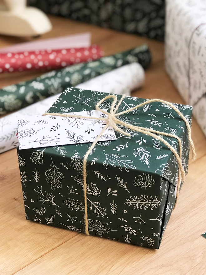 A gift wrapped in deep green wrapping paper with a Christmas botanical design lays on a wooden floor.