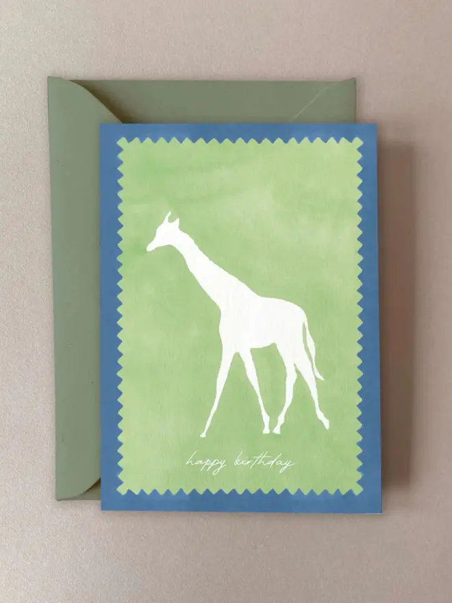 Birthday card featuring a white giraffe silhouette on a soft green background with a zigzag edge detail, bordered in blue and paired with a matching green envelope.