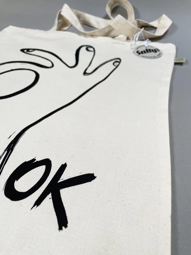 Close up of flat lay of Ok tote bag with black ink hand drawn in an OK gesture - Saltys swing tag tied to one handle and showing the fairtrade label