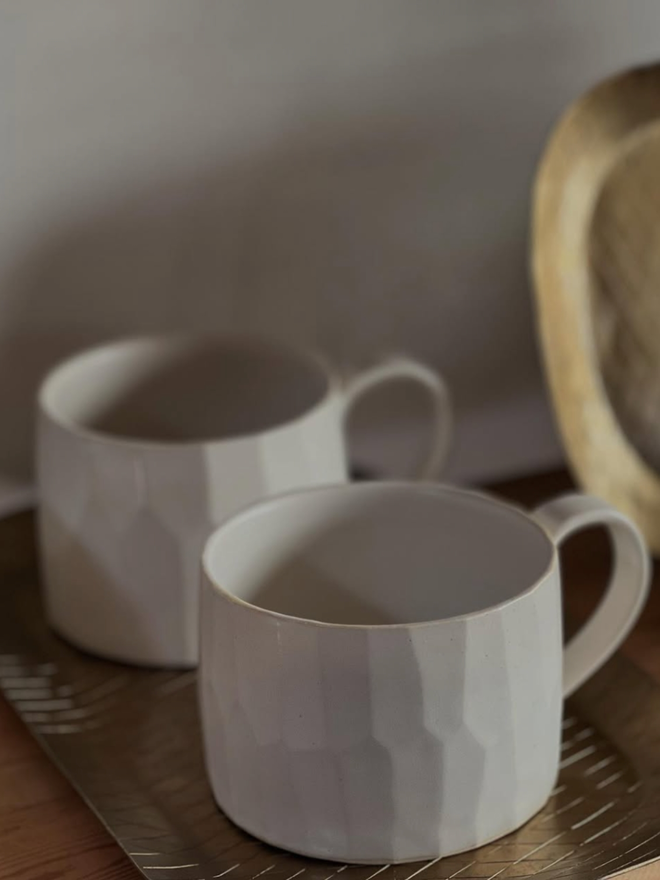faceted matte white ceramic mug. pair of mugs photographed on brass trays with grey wall in the background.