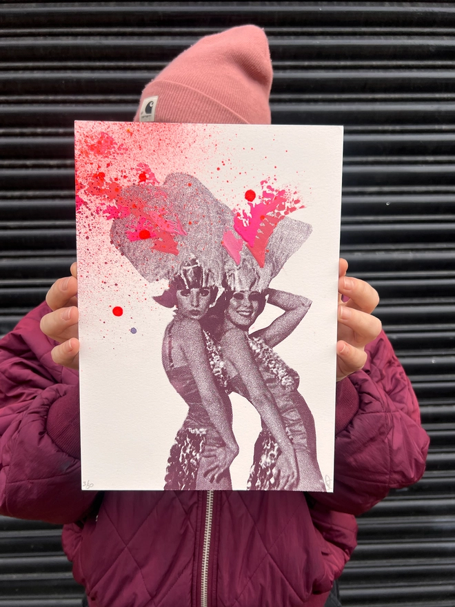 woman artist holding an A4 colourful pink and peach and burgundy print of 2 women in 1920s party clothes with hand painted swooshes and splats, 