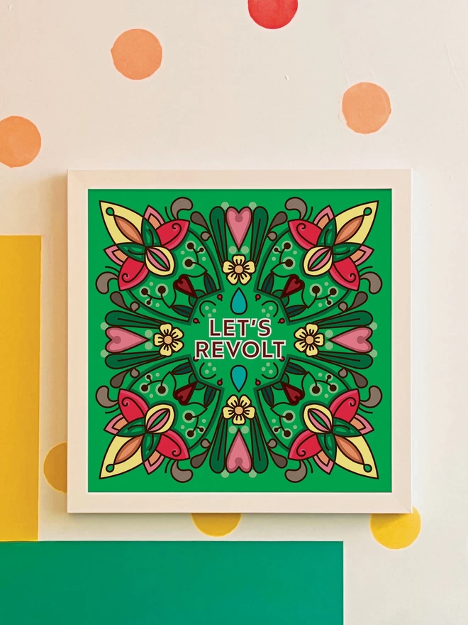 A symmetrical square design in greens, yellows and pinks with Let’s Revolt at the centre is hung in a white frame on a white wall with yellow, orange, green and blue spots, and a green and yellow rectangle painted in the bottom left hand corner. 