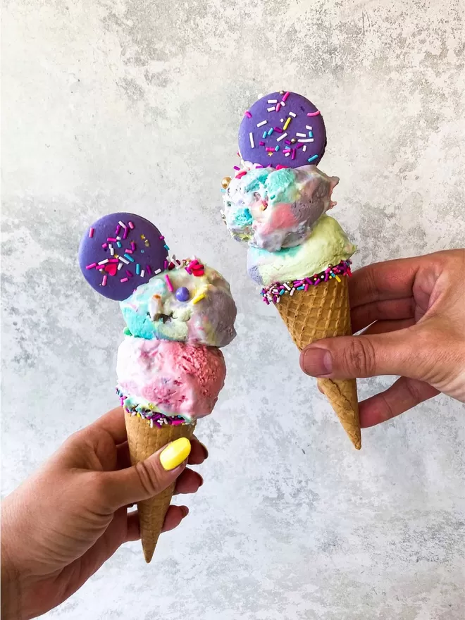 two hands holding two ice cream cones with colourful sprinkles, topped with a bright purple macaron
