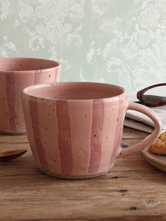 Handmade ceramic stoneware mug in pink stripe sat on a wooden surface with another mug in the same style sat behind it. The background also shows a newspaper with glasses on top, a brass spoon and a plate with cookies. 