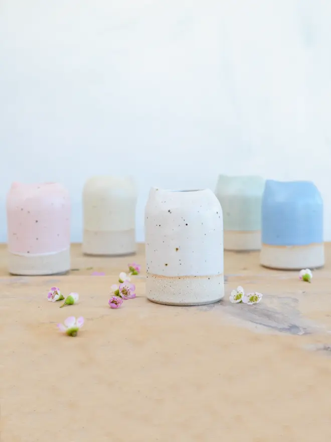 Five milk jugs in white, yellow, pink, blue and mint green on a wooden table surface dotted with little white and pink wildflower heads.