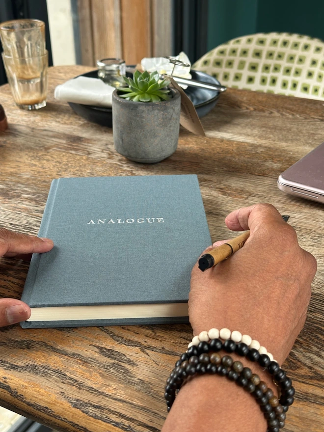 A notebook covered in grey linen and printed with 'Analogue' in gold lettering is held in a man's hands, along with a pen.