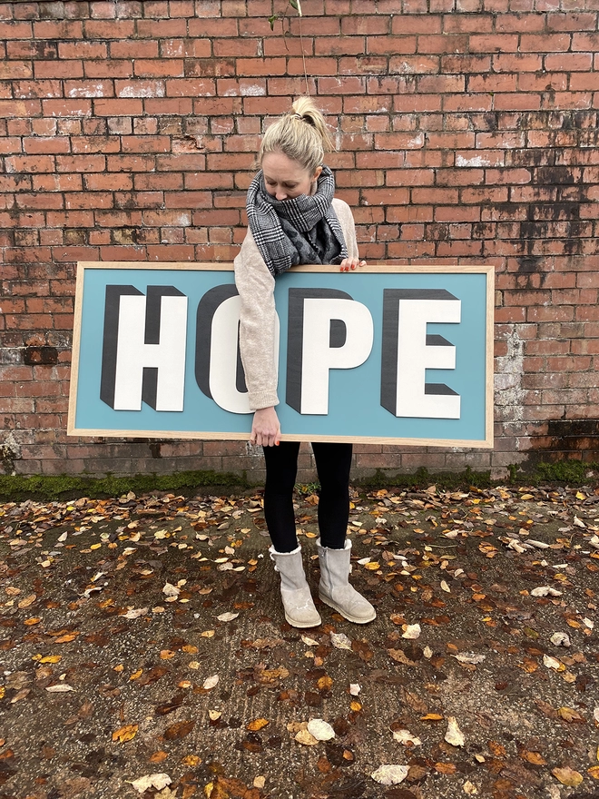 A person standing in front of a red brick wall holding a painted wooden sign which reads HOPE on a teal painted back board 