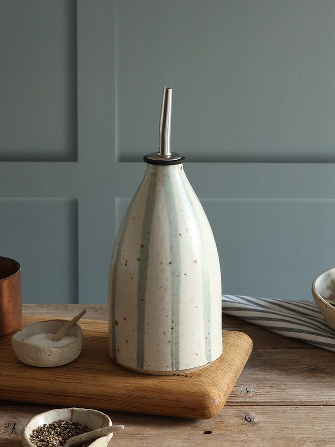 Handmade Ceramic Stoneware Oil Pourer in Blue and White Stripe sat on a wooden chopping board on top of a wooden surface next small handmade ceramic dishes containing salt and pepper, a brass measuring cup and a blue and white striped napkin. 
