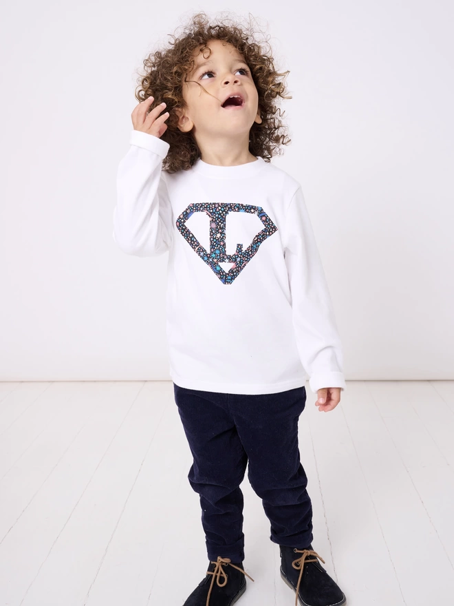 A boy wearing a white long sleeve t-shirt appliquéd with a superhero motif  featuring a letter L initial inside it. The appliqué is made from a starry Liberty print .
