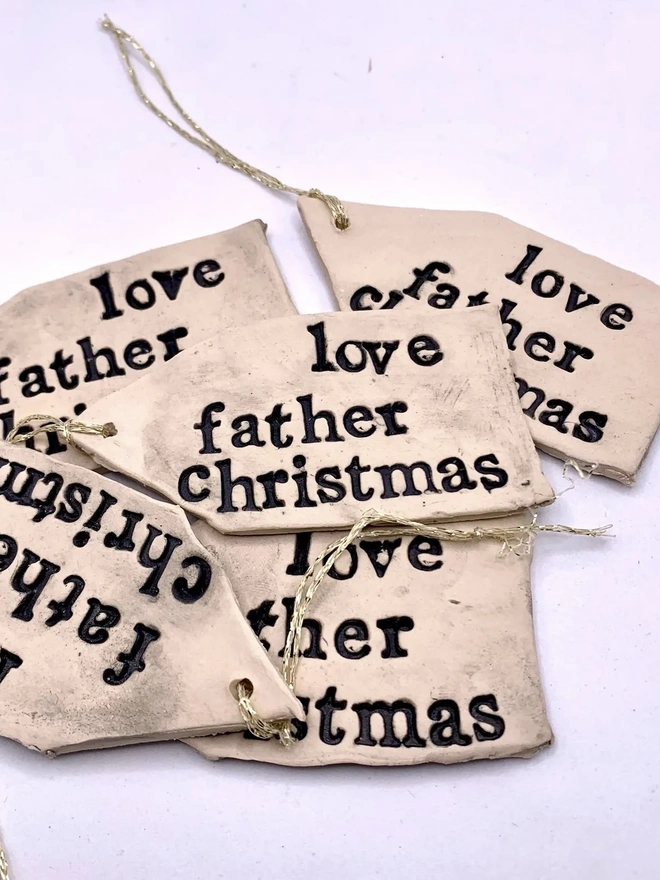 'Love Father Christmas' Handmade Ceramic Gift Tags, a group of ceramic gift tags attached sitting on a ale purple surface. 