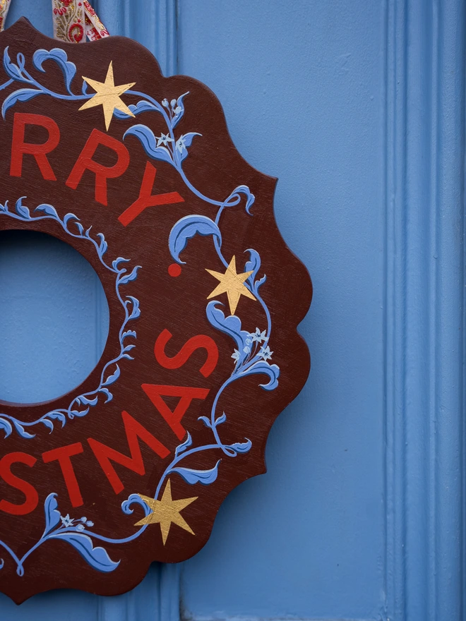 Depp Reddish Brown Hand painted Christmas Wreath with red lettering and gold leaf stars on a blue front door