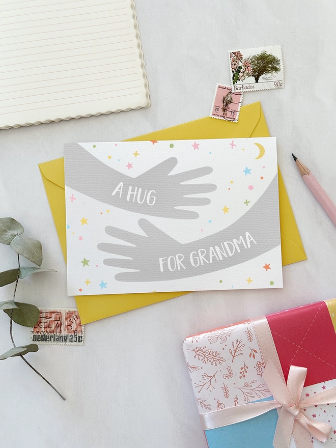 A friendship greetings card with a hugging arms design and personalised wording that reads "A hug from..." lays on a yellow envelope on a white desk.