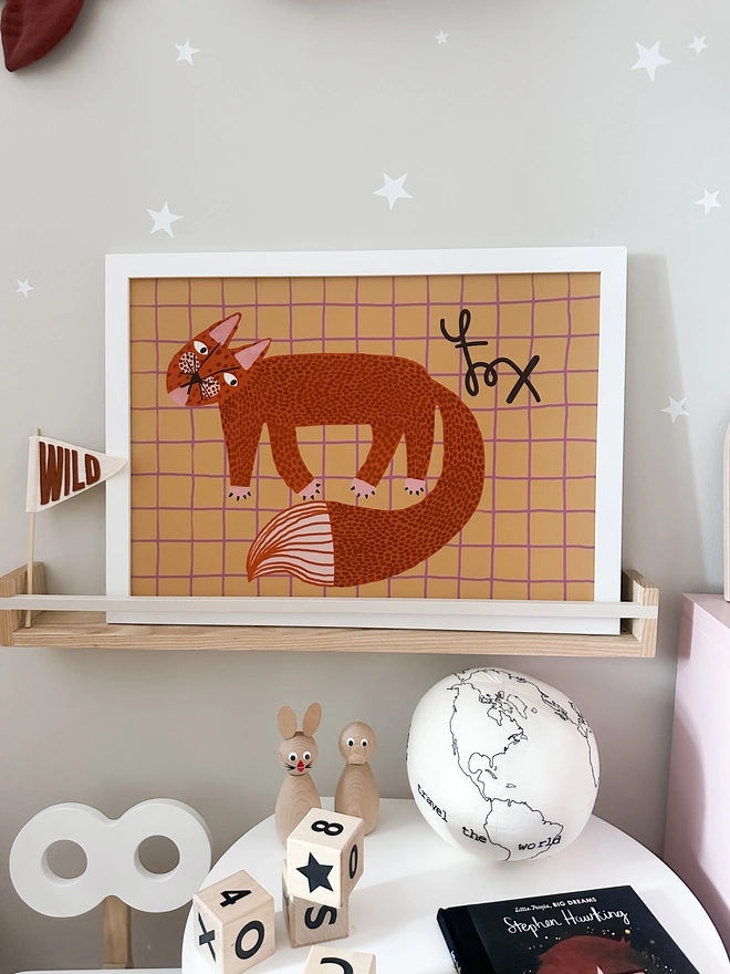 A quirky kids wall art print of a charming and fun fox. Styled propped up on a shelf in a Scandinavian style modern kids room or play room. 