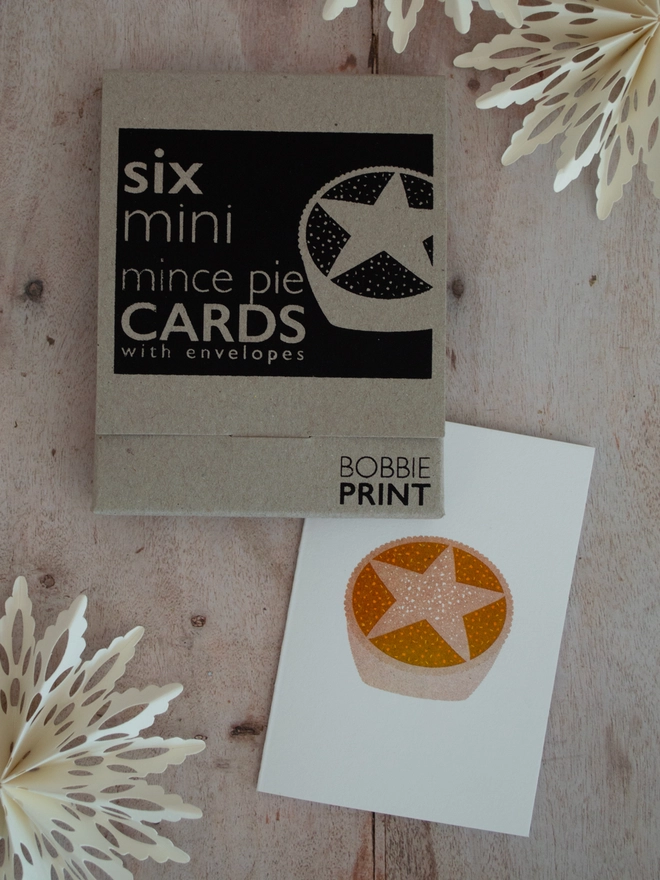 A closed box of six festive mince pie mini christmas cards lies on a wooden table, to the side of the box which is screen printed with a black label lies a small white card, risograph printed with a delightfully cute mince pie illustration.