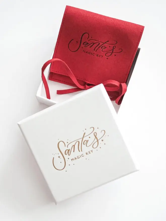 A white gift box and red suede pouch, both printed with the "Santa's Magic Key" logo. The pouch is tied with a thin red suede ribbon.