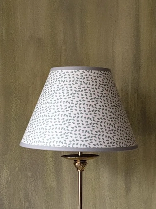 Paper Empire Lampshade. A tapered lampshade covered in a delicate grey and white scattered leaf print with a grey trim, shown on a brass lamp base. The subtle hand-printed pattern adds soft texture to a bedroom or sitting room.