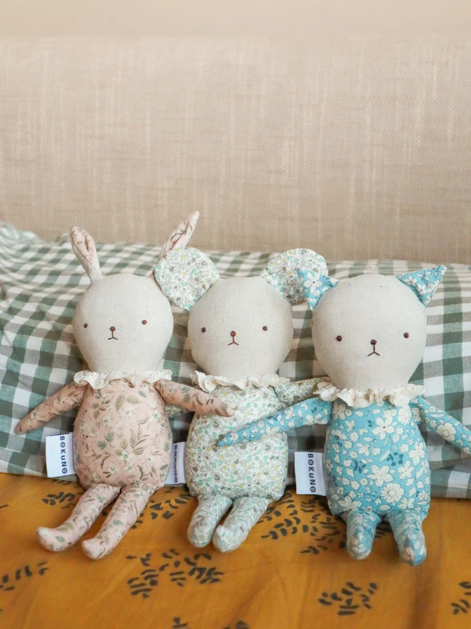 Three handmade fabric dolls with pastel floral prints: a cat, a mouse, and a rabbit. Each has a linen face, embroidered features, ruffled collars, and a 'BOKUNO' tag, highlighting their artisan craftsmanship. Perfect for gifting or collecting.