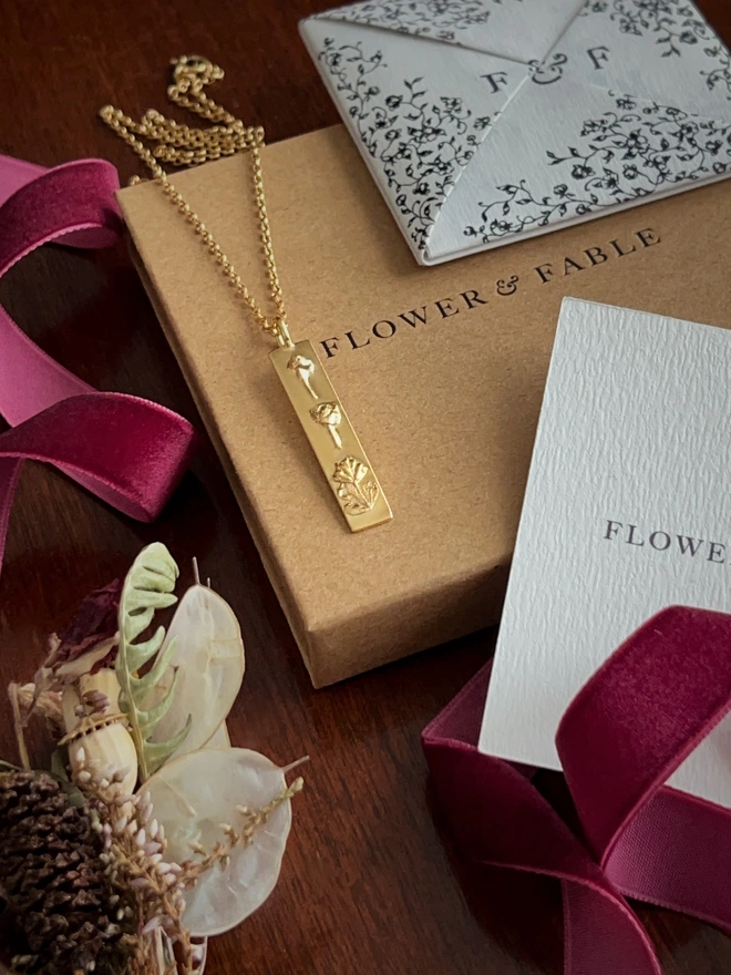Gold bar pendant belcher necklace representing luck draped over flower & fable packaging