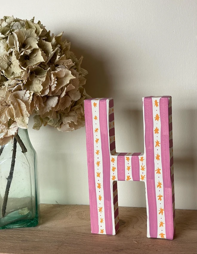 A hand-painted decorative initial letter  with stripes on a wooden table against a light wall. 