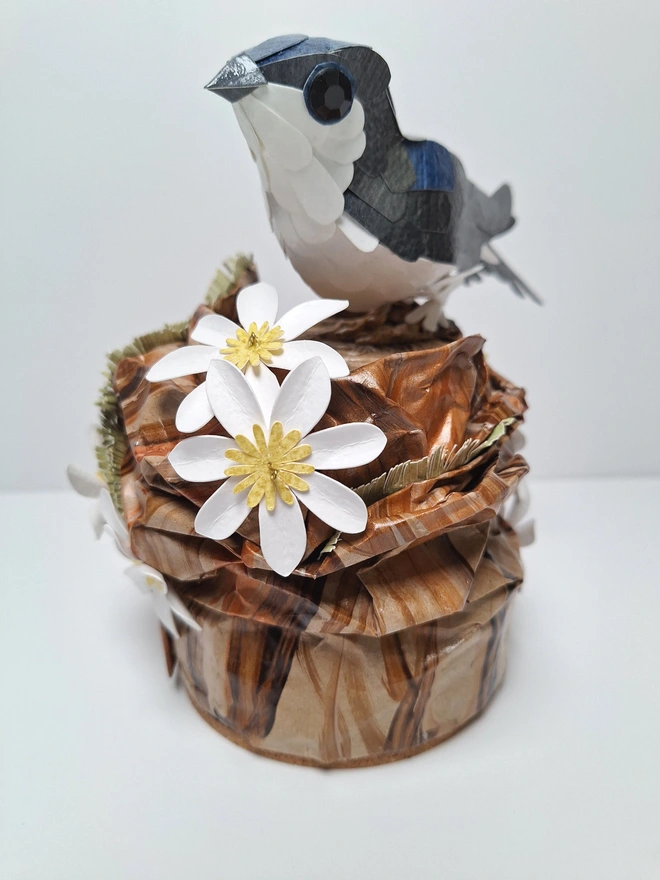 Housemartin bird sculpture, perched on a floral tree stump. This handmade sculpture is made from recycled paper. The bird is black and blue, with a white belly. White and yellow flowers adorn the tree stump. Front view.