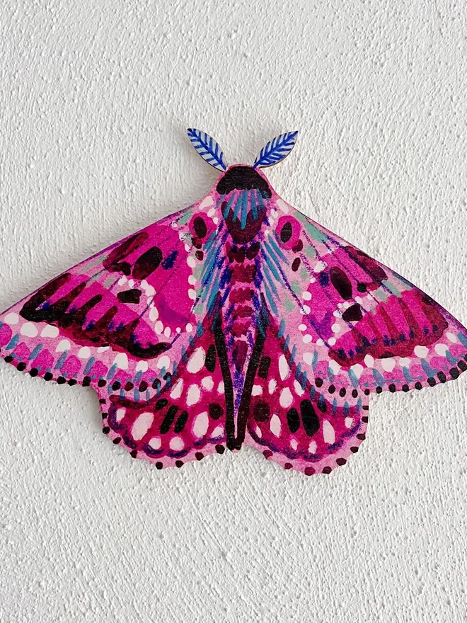 wooden moth wall decoration. pink wooden moth decoration hung on plain white wall.