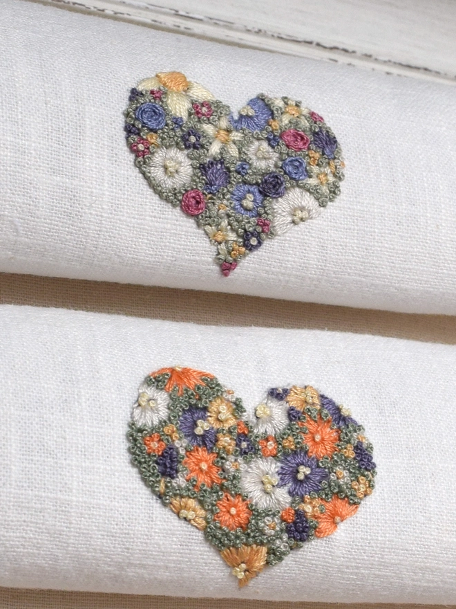 An embroidered Floral Meadow Heart, of Lavender Blues and Buttermilk yellow blossoms. Also, an embroidered Sunshine Garden Heart of Golden Yellows and Bright Orange Blossoms with Green French Knot grass background.  