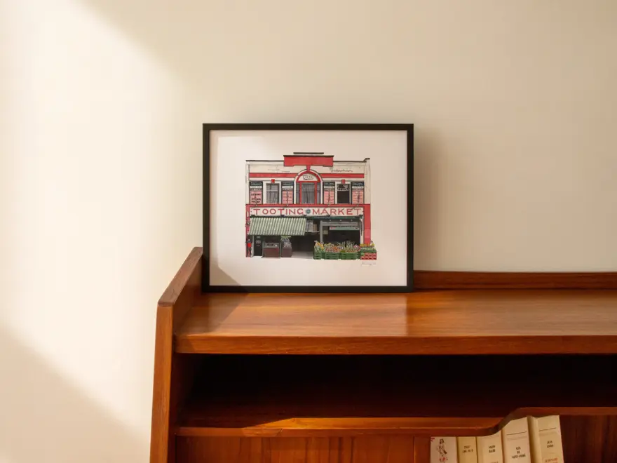 Watercolour illustration of Tooting Market, framed in black and sitting on a wooden desk against a cream wall