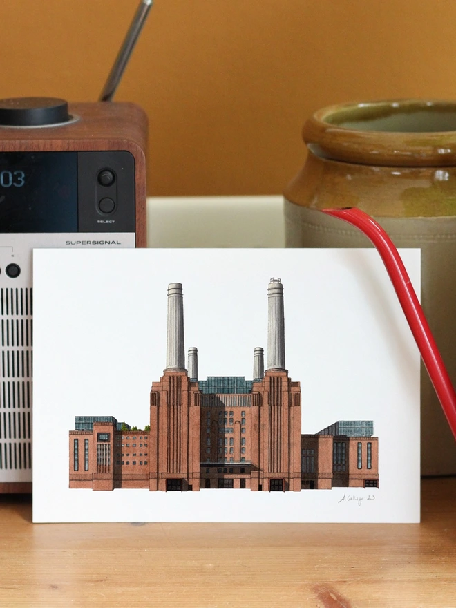 A detailed architectural illustration of Battersea Power Station on a print, displayed on a wooden surface alongside a digital radio and a red watering can.