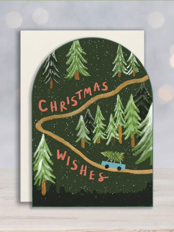 Arch shaped Christmas card showing a car going uphill carrying a Christmas tree, on a green background. Message reads Christmas Wishes