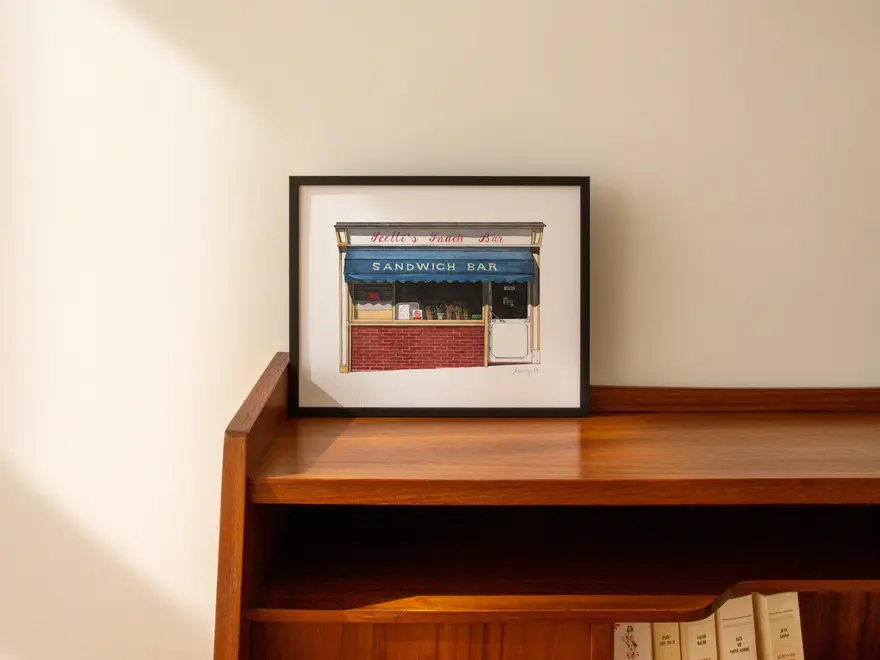 Clerkenwell Scotti's Snack Bar Giclée Print (Unframed), a print of a snack bar sitting in a black frame on a wooden surface against a plain backdrop. 
