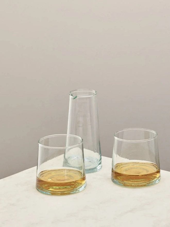 Tall Jug, a tall glass jug sitting on a plain counter top against a white backdrop. It is  accompanied by another smaller glasses. 
