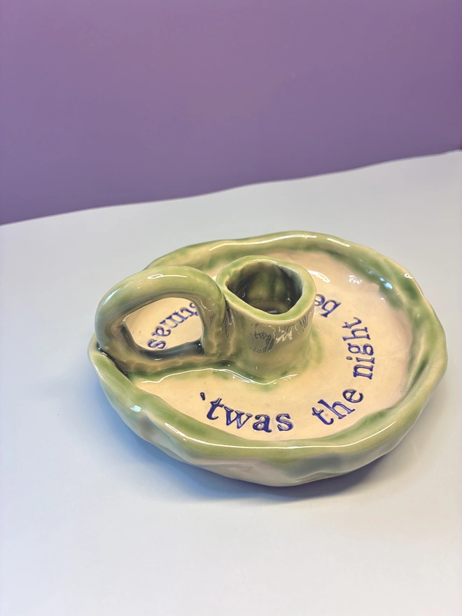 Handmade ceramic Christmas taper candlestick holder with a soft green glazed edge and the words “‘Twas the night before Christmas” in dark blue lettering, arranged around the candle holder opening. Photographed against a pale background.