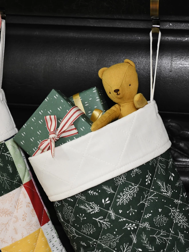 A quilted green stocking with an ivory turnover cuff hangs on a golden stocking hanger on a black mantlepiece. Small gifts and toys poke out of the top.