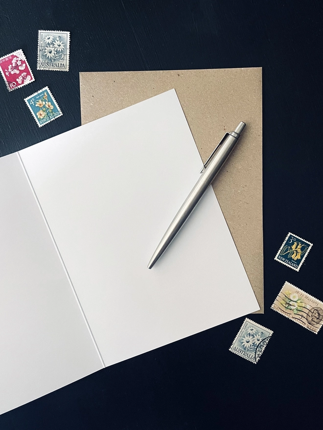 Open Greetings Card with Brown Kraft Envelope on Dark Charcoal Coloured Desk - Blank Interior - Floral Postage Stamps - Silver Parker Pen