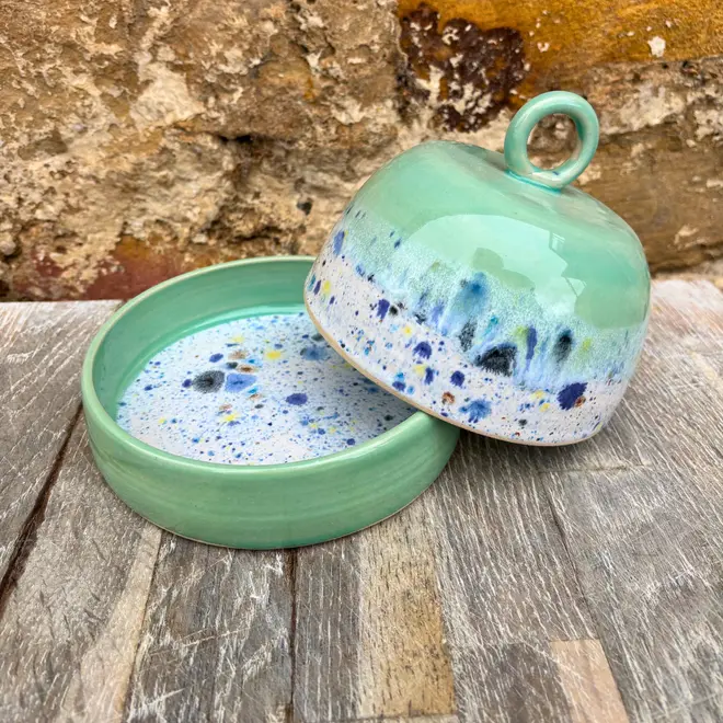 Shoreline Butter Dish, a colourful butter dish placed on a wooden surface against a brick wall. The lid is coming off in the image. 