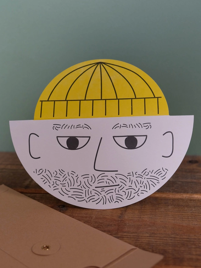 A round card with a male bearded face and yellow pop up hat. The card is hand letterpress printed in black and yellow ink on a light grey card. It is standing on a wooden shelf with green background.