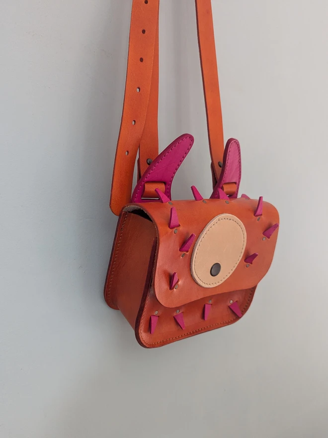 Orange leather monster cross body bag hanging against a blue wall, side view.