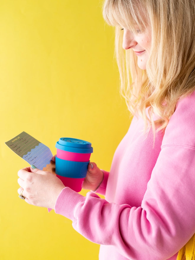 woman in pink jumper holds tear off shopping list in her hand with pink and blue coffee cup