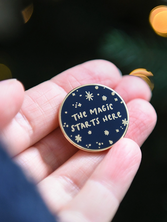 A navy blue and gold enamel pin badges with a starry design and the words "The magic starts here" rests on an open hand.